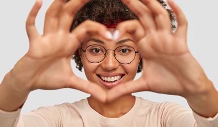 Photo of attractive young woman makes heart shape gesture over face, smiles broadly, demonstrates her love to boyfriend, wears round spectacles, isolated over white background, gestures indoor
